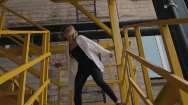 Siyah beyaz giyen havalı genç bir kadın sarı endüstriyel bir merdivende dans ediyor. Eğleniyor, kendine güveni tam ve hayatı dolu dolu yaşıyor..