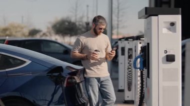 Orta yaşlı bir adam mavi elektrikli arabası şarj istasyonunda şarj olurken akıllı telefonunu kontrol ediyor. Giyinik ve elinde kahve fincanı var..