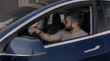 Odaklanmış bir erkek sürücü mavi bir arabada oturur, pencereden dışarı bakarken direksiyonu tutar, belli ki bir yolcuyu bekler..