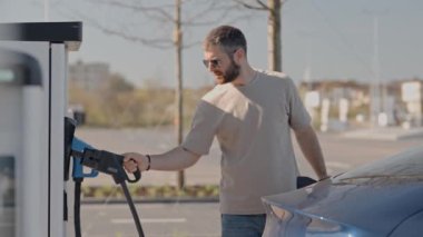 Orta yaşlı bir adam, güneşli bir günde mavi elektrikli arabasını modern bir şarj istasyonuna bağlıyor..