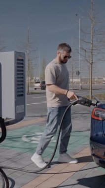Genç bir erkek, güneşli bir günde mavi elektrikli aracının şarj aletini tutarken şarj istasyonunda akıllı telefonunu kontrol eder..