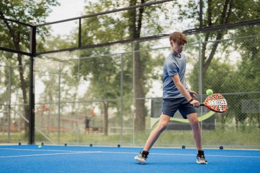 Açık hava mavi kortunda Padel tenis oynarken topa odaklanmış genç bir erkek oyuncu..
