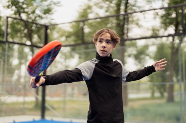 Siyah spor giyimli genç bir adam sahanın dışında Padel tenis oynuyor, oyuna odaklanıyor..