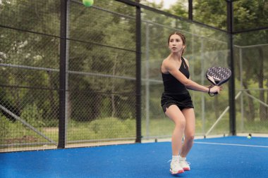 Açık havada tenis oynayan genç bir kadın topa vurmaya hazırlanıyor. Görüntü konsantrasyonu, hareketi ve atletizmi yakalar..