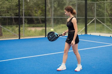 Mavi bir kortta Padel tenis antrenmanı yapan genç bir kadın. Spor giyim sektöründe ve oyuna odaklanmış durumda..