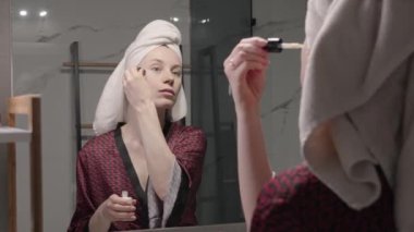 Banyodaki aynaya bakarken yüz kremi süren bir kadın. Evde bakım ve cilt bakımı rutini.