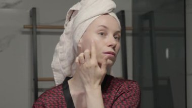 Banyoda kafasına havlu sarılmış bir kadın yüzüne yüz kremi sürüyor..