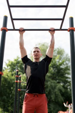 Dövmeleri olan odaklanmış genç bir adam güneşli bir günde açık hava spor salonunda barfiks çekerken görülüyor..