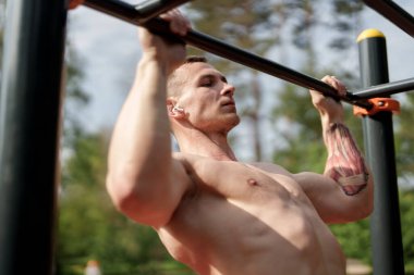 Görünür kasları olan atletik bir adam güneşli bir açık hava parkındaki barfiks çubuklarında çalışıyor. Doğanın içindeki gücü ve zindeliği gösteriyor..