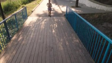 Parktaki ahşap bir köprüde koşan çekici genç bir kadın. Spor kıyafetleri giyiyor ve müzik dinliyor. Bu klip fitness, sağlık ve sağlık videoları için mükemmel..