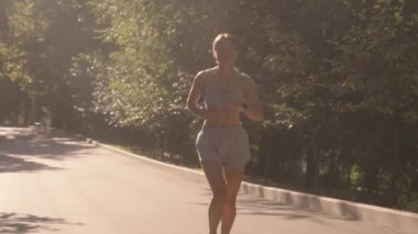 Genç bir kadın parkta sabah koşusundan zevk alır. O atletik giysi giyiyor ve kameraya doğru koşuyor. Güneş parlıyor ve ağaçlar yeşil.
