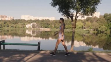 Güneşli bir günde genç bir kadın parkta koşuyor. Spor kıyafetleri giyiyor ve göl kenarında koşuyor. Bu video fitness, sağlık ve pozitif bir yaşam tarzını çağrıştırıyor..
