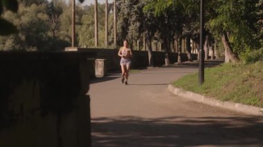 Genç bir kadın parktaki ağaçların arasında sabah koşusundan zevk alıyor. Spor kıyafetleri ve koşu ayakkabıları giyiyor..