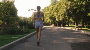 Genç bir kadın parktaki patikada kameradan uzaklaşıyor. Spor kıyafeti giyiyor ve yaz havasının tadını çıkarıyor..