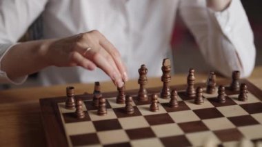 Beyaz gömlekli bir iş kadını ahşap bir masada oturuyor ve ev ortamında satranç oynuyor. Kral satranç taşını ileri taşıyor..