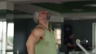 Orta yaşlı bir adam, modern bir spor salonunda yorucu bir çalışmadan sonra biraz dinlenip nefes alıyor..