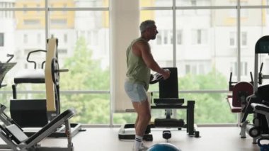 Modern bir spor salonunda ağırlık plakasıyla çalışan bir adam. Forma ve dayanıklılık antrenmanına odaklanıyor. Arkaplanda ekipman ve parlak pencereler.