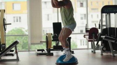 Spor salonunda çalışan bir adam, bir Bosu topunun üzerinde denge kurarken, bir çaydanlıkla çömelme hareketi yapıyor..