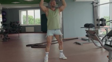 Şortlu ve kolsuz tişörtlü formda bir adam geniş ve iyi donanımlı bir spor salonunda halterli çömelmeler sergiliyor..