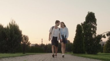 Birlikte parkta yürürken el ele tutuşan ve gülümseyen genç bir çift. Yazın gün batımında güzel bir gezintinin tadını çıkarıyorlar..
