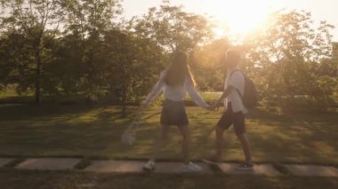 Gün batımında parkta yürüyen aşık genç bir çift. El ele tutuşup gülüyorlar. Arkalarında güneş parıldıyor..