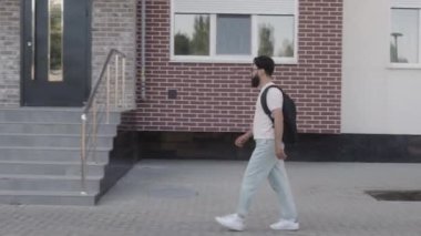 Beyaz gömlek, açık renk pantolon ve sırt çantalı gözlüklü genç bir adam tuğla bir binaya girmek için merdivenlerden çıkar..