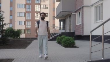 Gözlüklü mutlu sakallı bir adam şehrin dışında yürüyor. Gülümsüyor ve kameraya bakıyor..