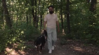 Gözlüklü ve sakallı bir adam, Bernese Dağ Köpeğiyle ormanda güneşli bir sabah yürüyüşünden zevk alır..