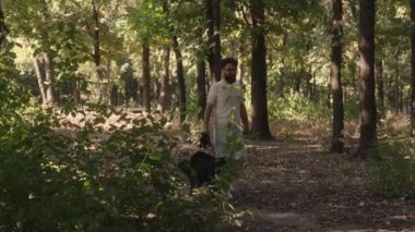 Bir adam köpeğini güneşli bir ormanda tasmayla gezdirir. Ağaçların arasında kirli bir yolda yürüyorlar.. 