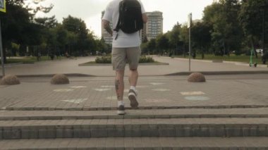 Dövmeli, beyaz tişörtlü, şortlu ve sırt çantalı genç bir adam şehir parkında kameradan uzaklaşıyor..