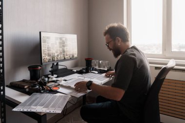 Odaklanmış bir fotoğrafçı, iyi aydınlatılmış bir odada, fotoğrafçılık ekipmanları ve büyük bir monitörle çevrili film negatiflerini analiz eder..