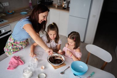 Anne ve iki genç kız mutfakta yemek pişirmenin keyfini çıkarıyorlar. Yemek pişirme sürecinde çikolata karışımı hazırlıyorlar..