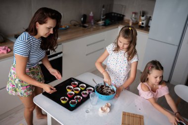 Bir anne ve iki kızı modern bir mutfakta kek pişiriyor. Sahnede aile bağları, takım çalışması ve yemek pişirme etkinliği sırasında eğlence anlatılıyor..