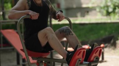 Açık havada kürek çekme makinesinde egzersiz yapan genç bir adam. Siyah bir atlet, siyah bir şort ve siyah bir beyzbol şapkası giyiyor. Güneşli bir günde spor yapıyor..