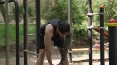Açık hava fitness parkında egzersiz öncesi esneme hareketleri yapan zinde bir genç adam. Antrenman kıyafetleri ve ters beyzbol şapkası giyiyor..