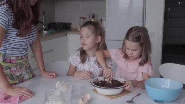 Anne ve iki kızı çikolatalı tatlı hazırlıyorlar. Beyaz bir mutfakta bileşenleri karıştırıyorlar. Aile kek veya kurabiye pişiriyor. Bu sıcaklık, bağlılık ve sevgiyi çağrıştırıyor..