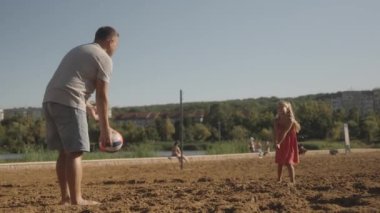 Bir baba ve küçük kızı, kumsalda voleybol oynuyorlar. Babası topa nazikçe vururken kız gülüyor ve eğleniyor.