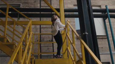 Şık genç bir kadın, sarı endüstriyel bir merdivende dans ediyor. Kişiliğini ve kendine olan güvenini bir şehir ortamında sergiliyor..