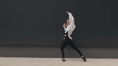Beyaz gömlekli ve siyah pantolonlu genç bir kadın şehir caddesinde zarif bir şekilde dans ediyor. Arkasındaki siyah duvar onun hareketlerine katıksız bir zıtlık sağlıyor, zarafetini ve çevikliğini vurguluyor..