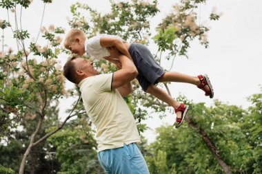 Baba, yaz günü yeşil bir parkta dışarıda oynarken oğlunu havaya kaldırıyor. İkisi de gülümsüyor ve birlikte vakit geçiriyorlar..