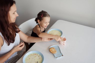 Bir anne ve küçük kızı parlak bir masada birlikte kahvaltı yapıyorlar. Kız mutlulukla çorba ve tost tabaklarının yanına uzanır.