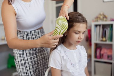 Bir anne rahat bir evde kızının saçını nazikçe tarar. Genç kız sakince oturur, basit beyaz bir gömlek giyer, şefkat ve şefkat duygusunu somutlaştırır.