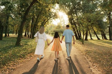 Bir aile, parkta güneşli bir yolda el ele yürüyor..