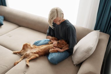 Sarı saçlı bir kadın kanepede oturup rahat köpeğini sevgiyle okşar. Aydınlık oda huzur ve sıcaklık yayıyor, huzurlu bir arkadaşlık ve neşe anı yakalıyor..