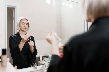 Siyah cüppeli bir kadın banyo aynasına bakarken serumu sürüyor. Sakinleştirici bir cilt bakımı rutini ve kişisel bakım anını gösteriyor..