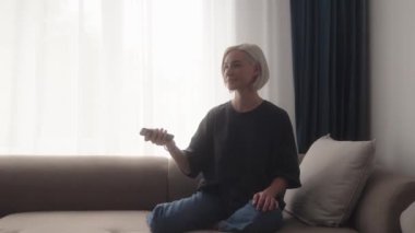 Oturma odasındaki kanepede oturan, rahatlayan ve televizyon izleyen genç bir kadın. Uzaktan kumanda kullanıyor, evde boş vaktinin tadını çıkarıyor..