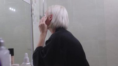 Kısa sarı saçlı genç bir kadın, banyoda aynanın önünde pamuk bir bezle yüzünü nazikçe temizliyor. Siyah bir bornoz giyiyor ve cilt bakımına odaklanıyor..