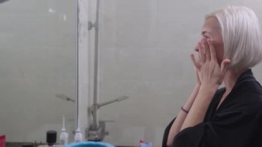 Kısa sarı saçlı, siyah bornozlu çekici bir kadın sabah duşundan sonra cilt kremi sürüyor. Güzellik ve cilt rutini.
