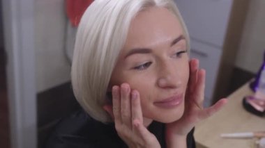 Genç sarışın bir kadın yüzüne nemlendirici sürüyor. Nazikçe cildine sürter. Siyah saten bir bornoz giyiyor ve yüzünde rahat bir ifade var..