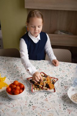 Okul üniformalı genç bir kız mutfak masasında meyve ve fındık içeren besleyici bir aperatif hazırlıyor. Görüntü bir bakım ve hazırlık hissi veriyor..